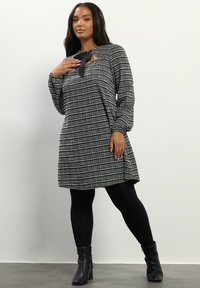 Schwarzes und weißes kariertes Kleid mit langen Ärmeln, Schlüsselloch-Ausschnitt und gebundenem Stoffdetail, kombiniert mit schwarzen Leggings und Stiefeln im Knöchelbereich.