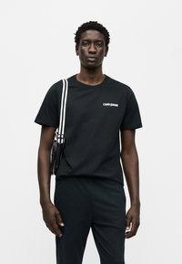 Μαύρο βαμβακερό t-shirt με λευκό λογότυπο "Converse" στο στήθος, σε συνδυασμό με μαύρο παντελόνι. Το μοντέλο φορά μια μικρή μαύρη τσάντα με ριγωτό λουράκι.