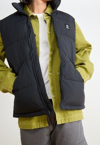 Czarna pikowana kamizelka z zamkiem błyskawicznym, przednimi kieszeniami i wysokim kołnierzem, noszona na zielonej koszuli na guziki z kontrastowym przeszyciem.