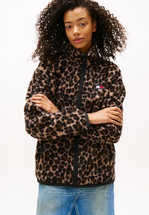Mujer con cabello rizado vistiendo una chaqueta de forro polar con estampado de leopardo y cremallera, con los brazos cruzados contra un fondo liso.