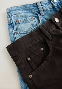 Jean en denim bleu plié partiellement recouvert par un jean brun foncé plié, montrant les poches avant avec des rivets visibles et des passants de ceinture.