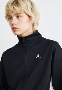 Svart zip-up jacka med hög krage, prydd med en grå Jordan-logga på vänster sida. Materialet verkar vara slätt och stretchigt.