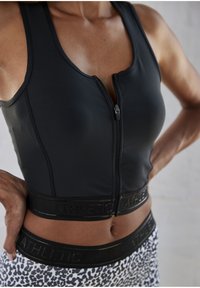 Black athletic crop top with a front zipper and black branded elastic band. Features smooth, fitted fabric and wide straps. Patterned leggings beneath.