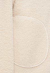 Κοντινό πλάνο από απαλό υλικό φλις σε κρεμ χρώμα με στρογγυλή αναδιπλωμένη τσέπη περιγραμμένη από λεία επένδυση.