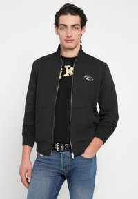 Giacca nera con cerniera e colletto alla coreana, con un piccolo logo sul lato sinistro. Indossata sopra una t-shirt nera con grafica e jeans blu.