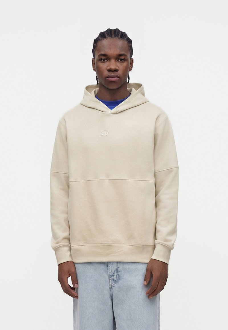 Ung mand med flettet hår iført en beige Nike-hoodie over en blå skjorte og lyseblå jeans, stående foran en hvid baggrund.