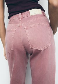 Roze traperice s medijem strukom, klasičnim dizajnom s pet džepova i vidljivom oznakom branda na pojasu.