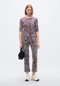 Σετ κορυφής και παντελονιών με λεοπάρ μοτίβο σε καφέ και κρεμ χρώματα. Το ύφασμα έχει απαλή υφή. Το μοντέλο κρατά μια μικρή μαύρη τσάντα και φορά κρεμ αθλητικά παπούτσια.