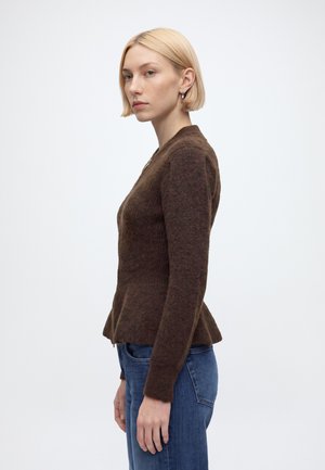 Jeune femme aux cheveux blonds courts portant un pull marron à manches longues et un jean bleu, debout de profil sur un fond blanc uni.