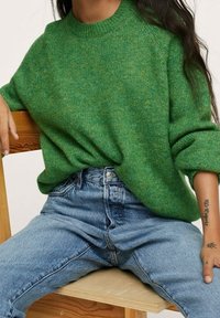 Grüner Strickpullover mit rundem Ausschnitt und Raglanärmeln, kombiniert mit hellblauen, hoch taillierten Jeans. Holzstuhl im Hintergrund.