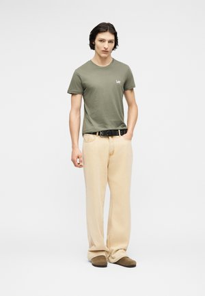 Junger Mann mit mittellangen dunklen Haaren, der ein olivgrünes Lee-T-Shirt, beige Hosen mit weitem Bein, einen schwarzen Gürtel und braune Schuhe trägt und aufrecht steht.