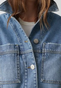 Džínová bunda v světle modré barvě se zvýšeným límcem, dvěma předními kapsami, stříbrnými druky a opotřebovanými okraji pro texturu.