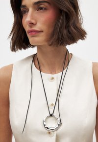 Collier avec pendentif en métal argenté sur un cordon noir, présentant une forme circulaire organique et texturée, porté sur un haut sans manches crème.