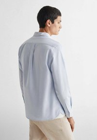 Lichtblauwe overhemdd met een rugplooi, lange mouwen en manchetten; gemaakt van zachte stof met een gladde textuur.