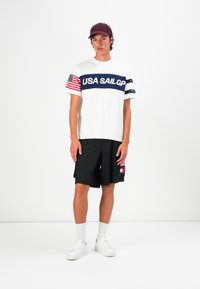 Tricou alb cu text albastru "USA SAILGP" și design cu steagul american; pantaloni scurți negri cu buzunar lateral, adidași albi și o șapcă cărămizie.