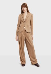 Kamel kaput od vune s jednim dugmetom, izrezanim reverima i dva prednja džepa, uparen s odgovarajućim širokim hlačama. Glatka tekstura.