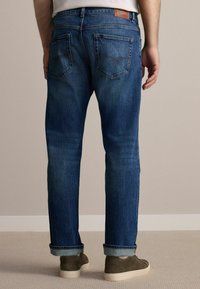 Jeans din denim de culoare albastră închisă, cu un finisaj decolorat, croială dreaptă, cinci buzunare și un patch din piele maro pe spate. Manșete răsucite și pantofi bej.