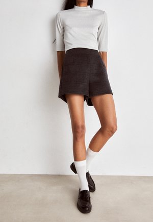 Frau trägt ein hellgraues Kurzarmshirt und dunkelgraue hochtaillierte Shorts, weiße Socken und schwarze Loafer, steht vor einer schlichten Wand.