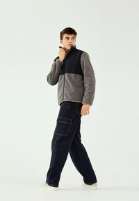 Chaqueta de forro polar gris y negra con cremallera y cuello alto. Pantalones cargo navy de corte holgado con bolsillos laterales y costuras blancas. Fondo claro.