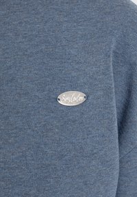 Tissu tricoté bleu avec une petite étiquette métallique ovale argentée attachée, gravée du mot "Smalotox."
