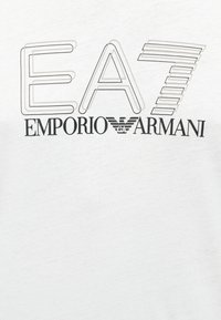 Άσπρο βαμβακερό t-shirt με έντονο μαύρο κείμενο: "EA7 EMPORIO ARMANI," με καλλιτεχνικά γράμματα και λογότυπο στο κέντρο.