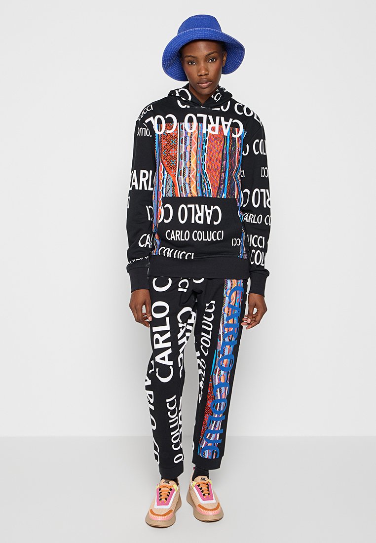 Sudadera y pantalones negros con patrones coloridos y texto blanco repetido. Un sombrero de cubo azul completa el atuendo, combinado con zapatillas de deporte en tonos pastel.