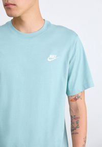 Άνδρας με μπλε αθλητικό T-shirt της Nike, που δείχνει μερικώς το πρόσωπό του και έχει τατουάζ στο δεξί του χέρι, σε λευκό φόντο.
