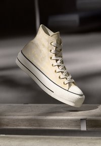 Ψηλό αθλητικό παπούτσι σε ανοιχτό κίτρινο χρώμα με υφασμάτινη υφή, λευκές κορδόνες και καουτσούκ σόλα που διαθέτει μια μαύρη ταινία στη βάση.