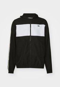 Svart zip-up jacka med vit panel över bröstet, med en huva och elastiska ärmslut. Lacoste-logotyp på bröstet och varumärke längs ärmarna.