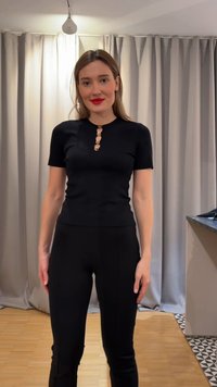 Naine seisab siseruumis, kandes seljas musta, kuldsete nuppudega, lühikeste varrukatega tihedalt istuvat topi ja musti pükse, taustal beežid kardinad ja puidust põrand.