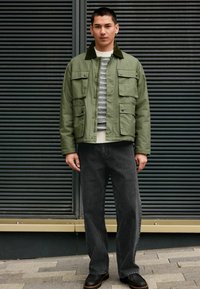 Olivenfarbene Jacke mit großen Klappentaschen, gestepptem Stoff und dunklem Kragen, kombiniert mit grau gestreiften Hosen und schwarzen Schuhen.