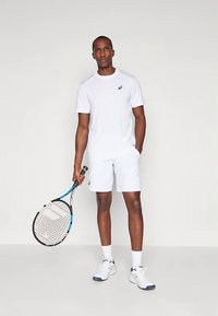 Valge sportlik T-särk ja lühikesed püksid hingavast kangast. Modell hoiab tennise reketit, jalas valged tossud siniste ja mustade detailidega.