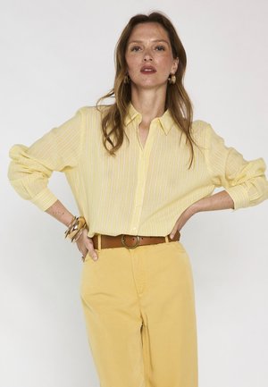 Femme debout, les mains sur les hanches, portant une chemise rayée jaune, un pantalon jaune, une ceinture marron, des boucles d’oreilles dorées et des bracelets, sur fond uni.