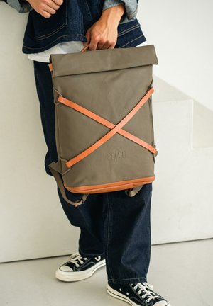 Person trägt dunkle Jeans und hält einen taupefarbenen und hellbraunen Rucksack mit gekreuzten Lederriemen und geprägtem Logo, steht vor einer hellen Wand.