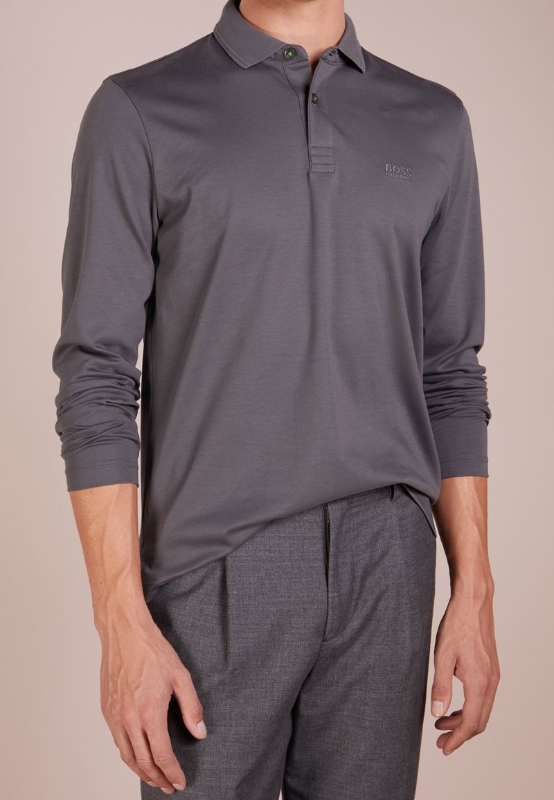 Szara koszulka polo z długim rękawem i kołnierzykiem, zdobiona subtelnym logo oraz zielonymi guzikami; zestawiona z szarymi, strukturalnymi spodniami.