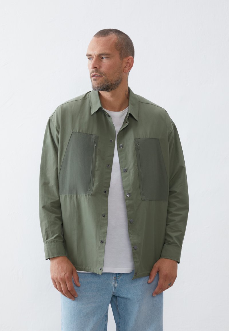 Olive green jacket with two large front pockets, snap button closure, and a relaxed fit. Worn over a white shirt and paired with light jeans.