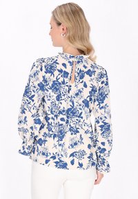 Blusa floreale con sfondo crema e motivi di rose blu, maniche a palloncino e apertura a chiave sul retro. Tessuto morbido.