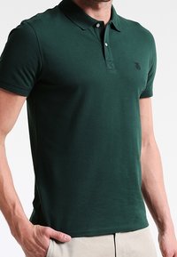 Green short-sleeve polo shirt made of cotton. Features a fold-down collar, three-button placket, and a small embroidered logo on the chest.