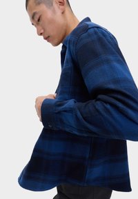 Mężczyzna zapina guziki na niebiesko-czarną koszulę w kratę z długim rękawem na białym tle.