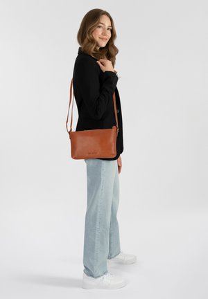 Braune Leder-Crossbody-Tasche mit glatter Textur, Reißverschluss und verstellbarem Riemen, präsentiert an einem Modell, das einen schwarzen Blazer und helle Jeans trägt.