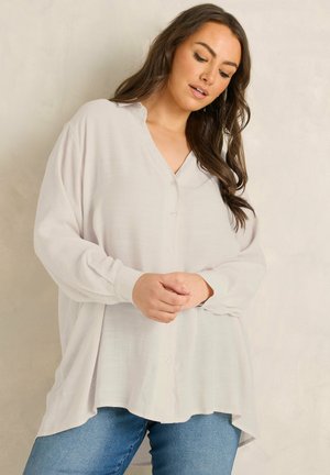 Mujer con cabello largo y oscuro, que lleva una blusa blanca suelta con botones y vaqueros azules, de pie frente a un fondo neutral y claro.
