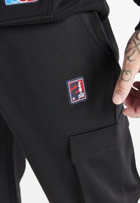 Nærbillede af sorte strukturerede bukser med et rødt, hvidt og blåt Looney Tunes "2" patch og en tatoveret hånd nær lommen.