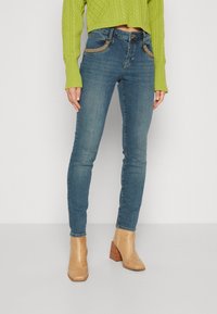 Jeans ajustados azules con costuras decorativas en los bolsillos, combinados con un suéter corto verde brillante y botas de tobillo de color marrón claro y textura.