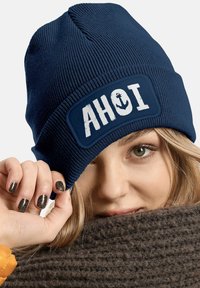 Navy blue knitted beanie with a white patch featuring the text "AHOI" and an anchor design. Texture appears ribbed and soft.