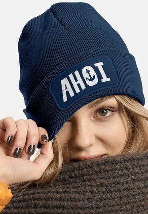 Navy blue knitted beanie with a white patch featuring the text "AHOI" and an anchor design. Texture appears ribbed and soft.