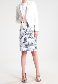 Blazer blanc sur un chemisier clair, associé à une jupe arrivant au genou avec un motif de feuilles tropicales bleues et des sandales à talons multicolores.