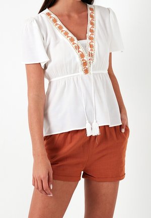Vit blus med kort pösiga ärmar, V-ringning, orange blommig brodyr och vita tofsar; kombinerad med rostfärgade shorts.
