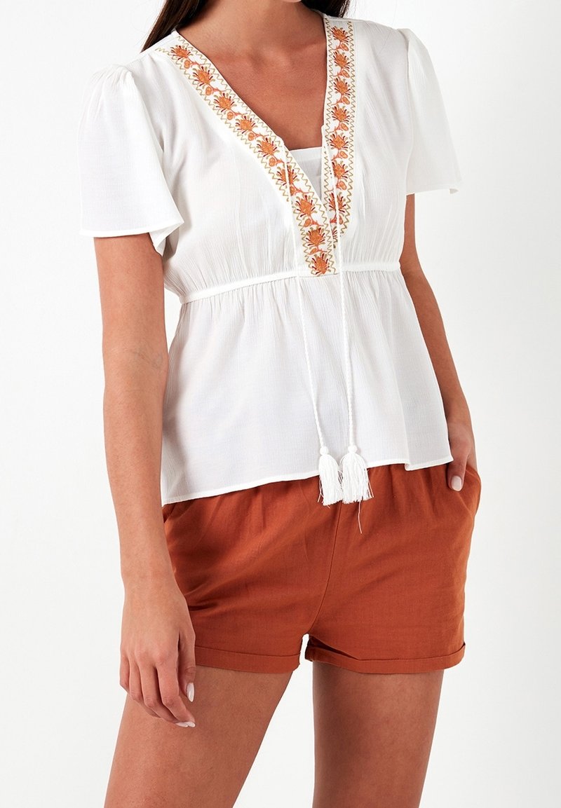 Blusa bianca con maniche a sbuffo corte, scollo a V, ricamo floreale arancione e frange bianche; abbinata a pantaloni corti color ruggine.