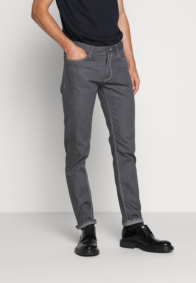 Man som bär tajta grå jeans och svarta läderskor, med ena handen i fickan och svart skjorta instoppad.