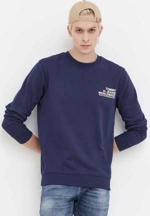 Felpa blu navale con polsini e orlo a coste, presenta un logo bianco sul petto. Indossata con jeans azzurri, su uno sfondo bianco.